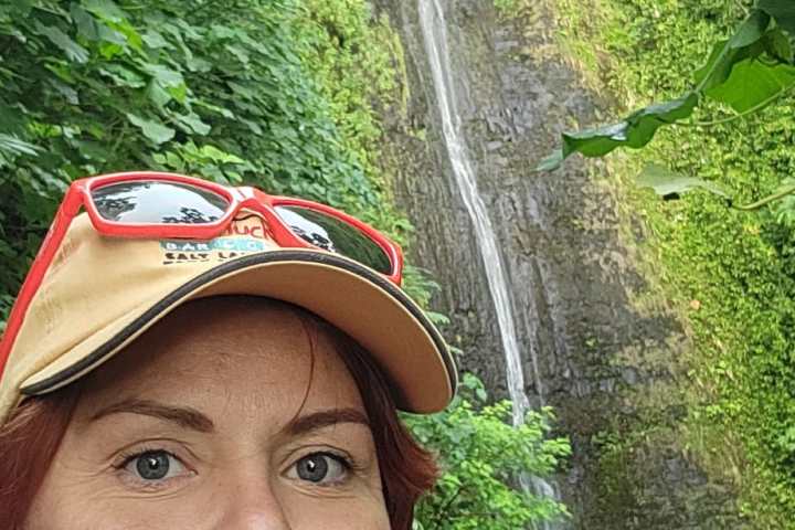 Person posing in front of a waterfall in a lush, green forest setting with a group of people nearby.